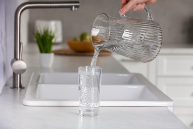 Woman pouring water from jug into glass at white countertop in kitchen, closeup
