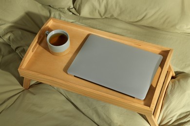 Photo of Wooden tray with modern laptop and cup of aromatic tea on bed indoors, above view