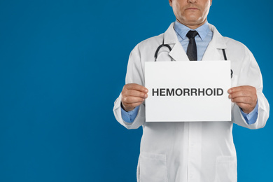 Photo of Doctor holding sign with word HEMORRHOID on blue background, closeup. Space for text
