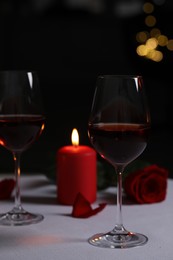 Photo of Glasses of red wine, rose flower and burning candle on white table against blurred lights. Romantic atmosphere