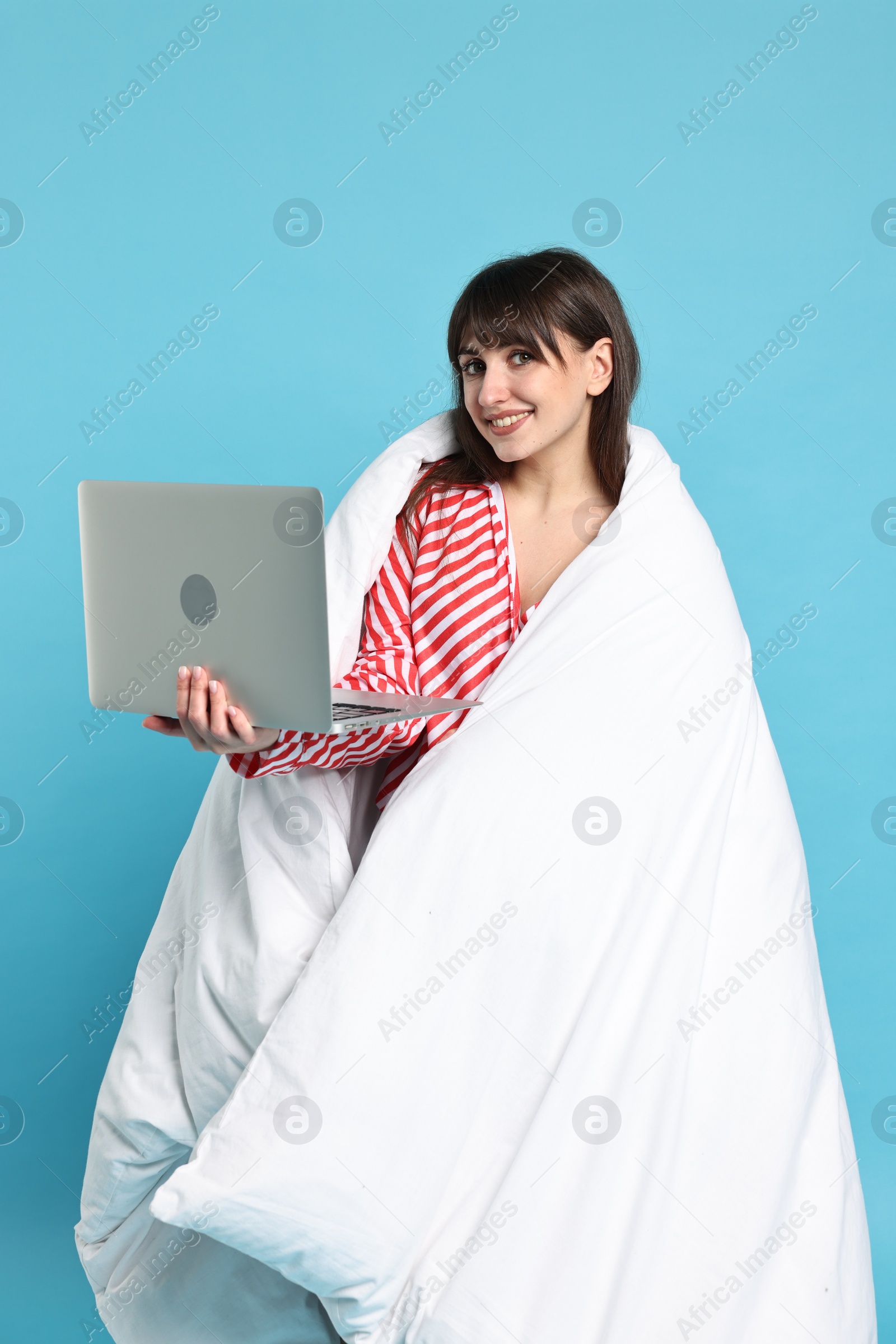 Photo of Happy woman in pyjama wrapped in blanket holding laptop on light blue background