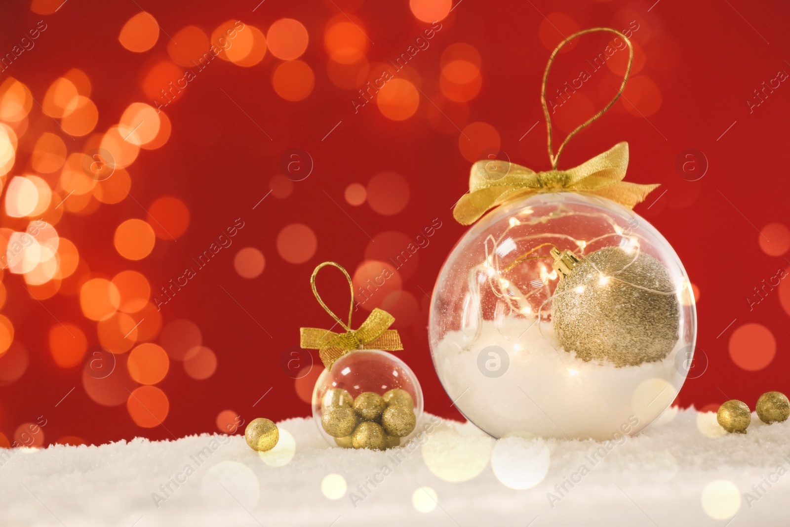 Image of Beautiful transparent Christmas ornaments with golden bows on snow against red background, bokeh effect. Space for text