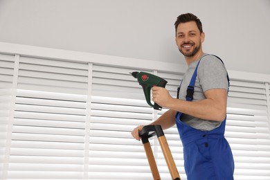 Photo of Worker in uniform installing window blind on stepladder indoors