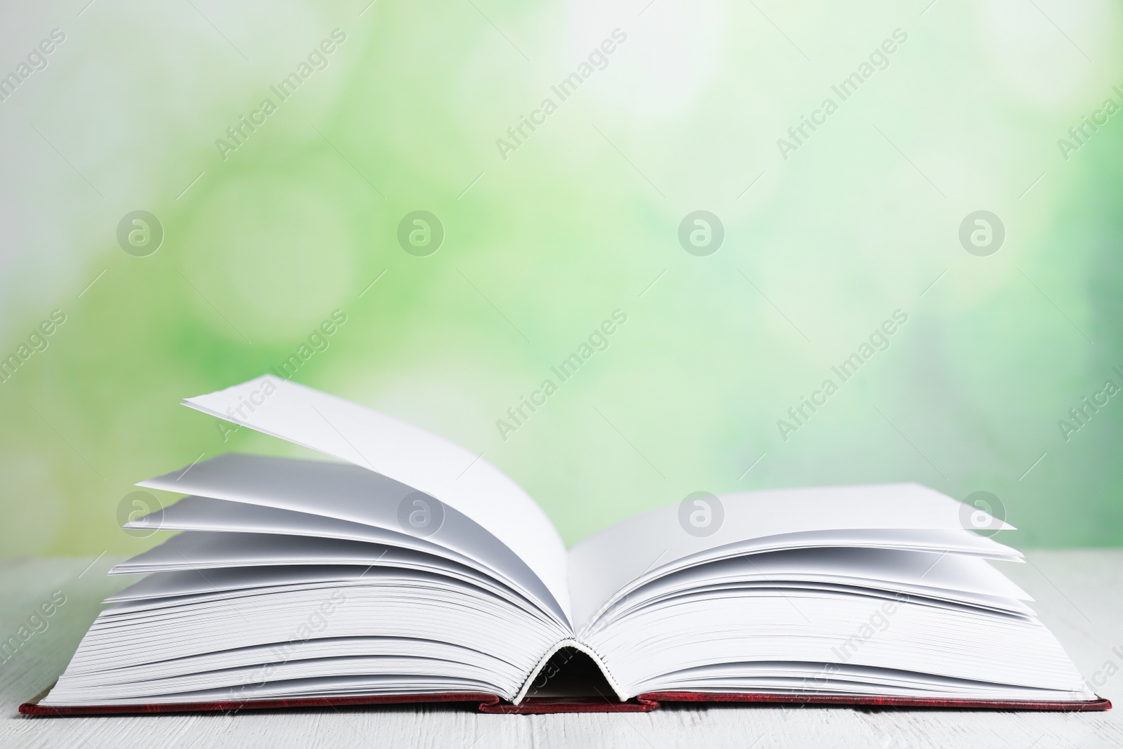 Photo of Open book on white wooden table against blurred green background. Space for text