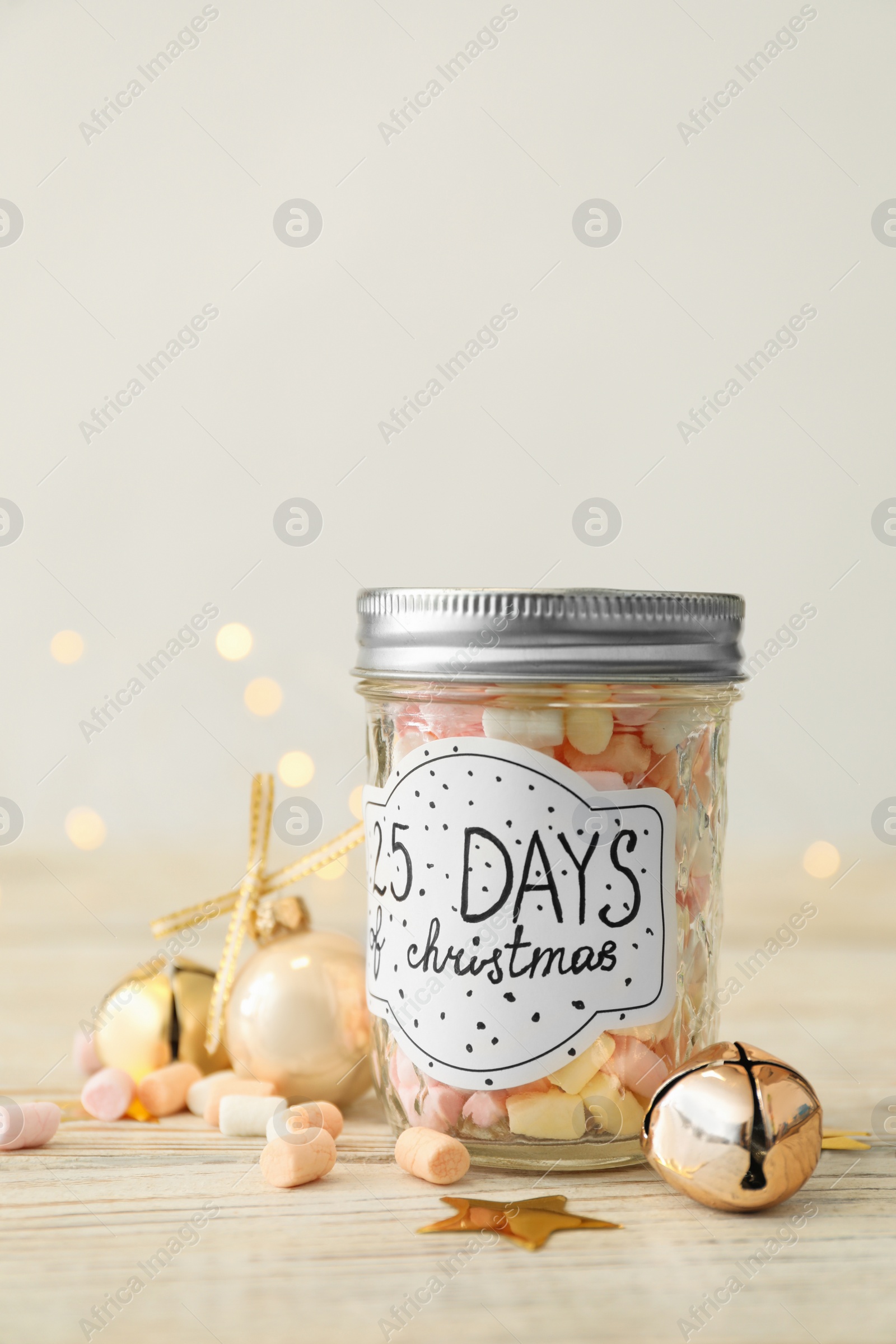 Photo of Glass jar with label 25 Days of Christmas and tasty marshmallows on white wooden table against blurred background. Advent calendar