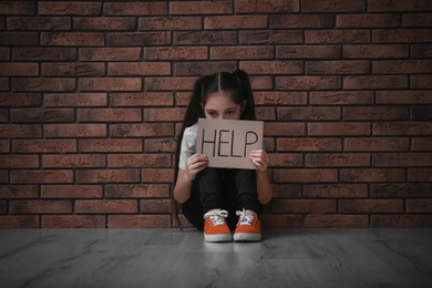 Photo of Sad little girl with sign HELP on floor near brick wall. Child in danger
