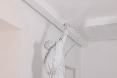 Photo of Decorator in protective overalls painting ceiling with spray gun indoors