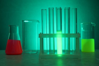 Photo of Laboratory glassware with luminous liquids on table against green background