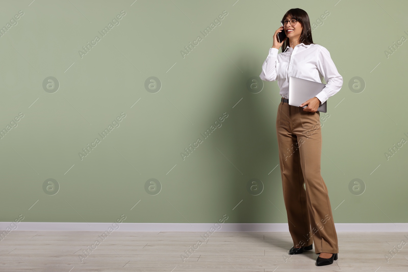 Photo of Happy secretary with laptop talking on smartphone near light olive wall indoors. Space for text