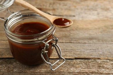 Photo of Tasty barbeque sauce in jar and spoon on wooden table, closeup. Space for text