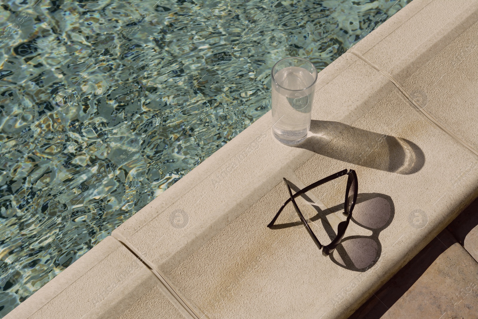 Photo of Stylish sunglasses and glass of water near outdoor swimming pool on sunny day, space for text
