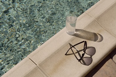 Stylish sunglasses and glass of water near outdoor swimming pool on sunny day, space for text