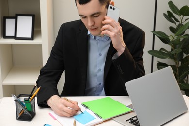 Photo of Man taking notes while talking on smartphone at table in office