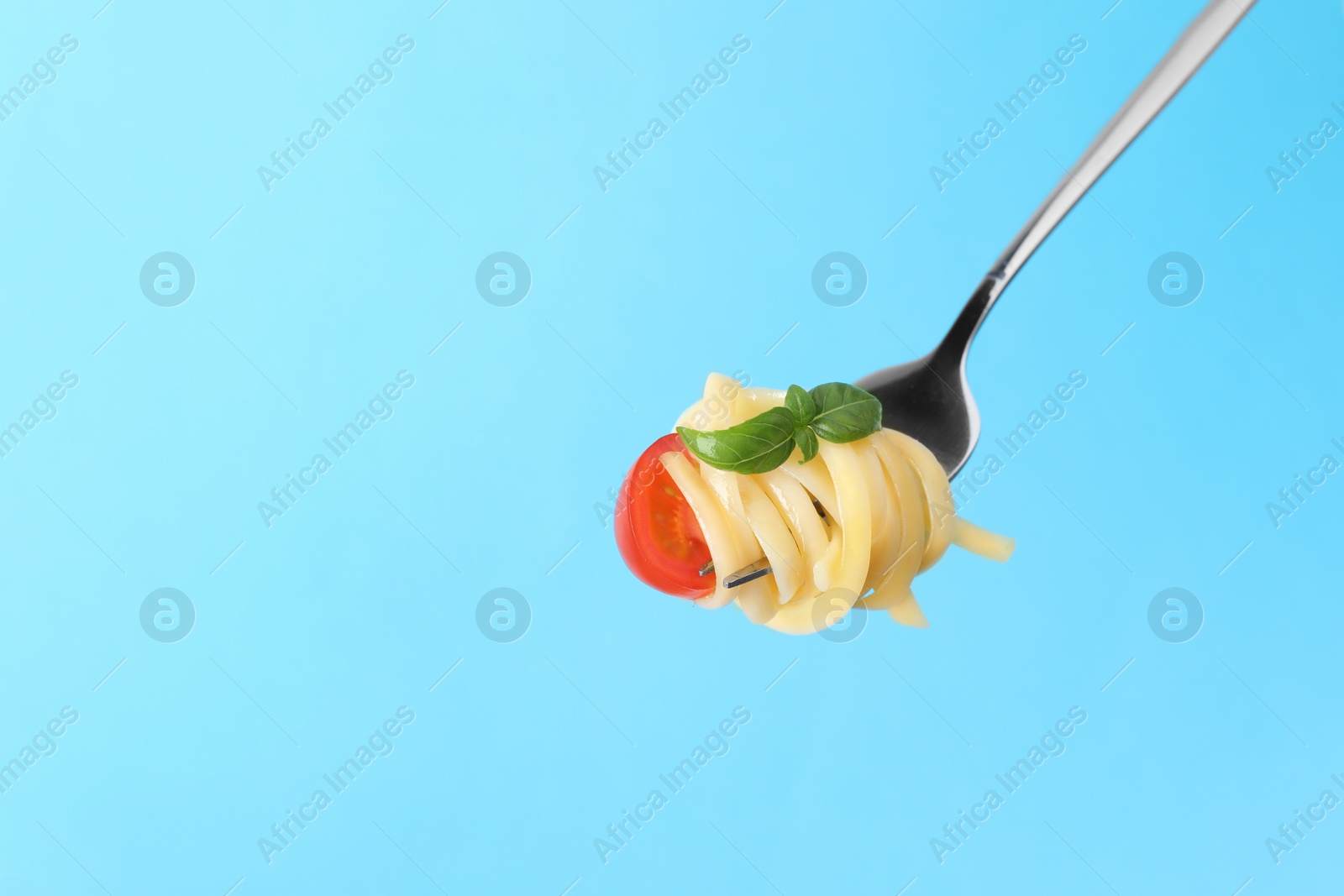 Photo of Fork with tasty pasta, basil and tomato on light blue background, space for text