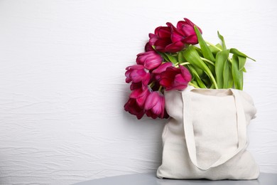 Tote bag with beautiful purple tulips on table near white textured wall, space for text