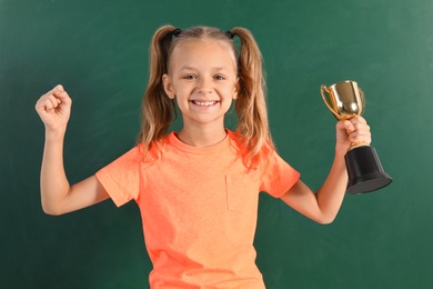 Photo of Happy girl with golden winning cup near chalkboard