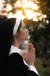 Photo of Young nun with hands clasped together praying outdoors on sunny day