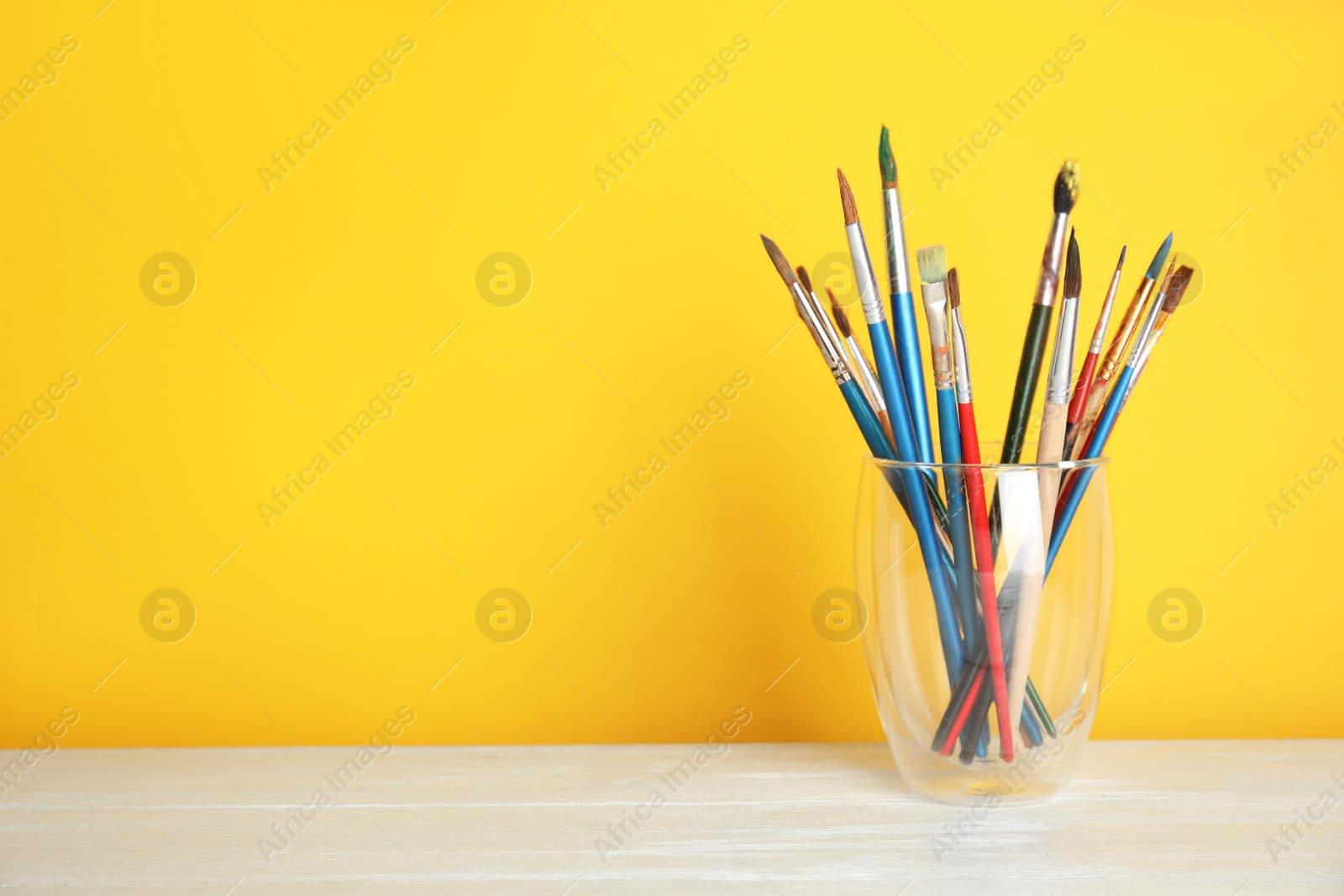 Photo of Glass with paint brushes on table against color background. Space for text