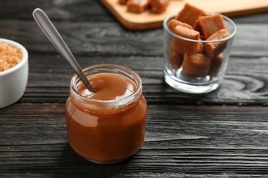 Jar with tasty caramel sauce and spoon on wooden table. Space for text