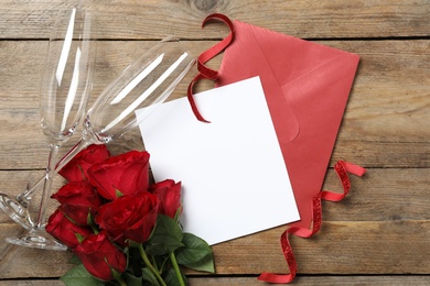 Photo of Flat lay composition with blank greeting card, glasses and roses on wooden table. Valentine's day celebration