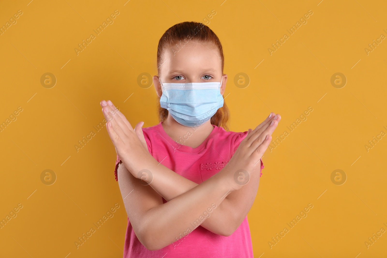 Photo of Little girl in protective mask showing stop gesture on yellow background. Prevent spreading of coronavirus