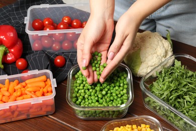 Woman putting green peas into glass container at wooden table, closeup. Food storage