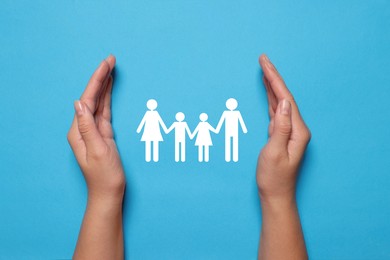 Image of Woman holding hands around paper silhouette of family on light blue background, top view. Insurance concept