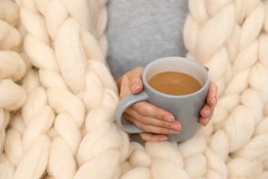 Woman wrapped in soft knitted blanket holding cup of coffee, closeup
