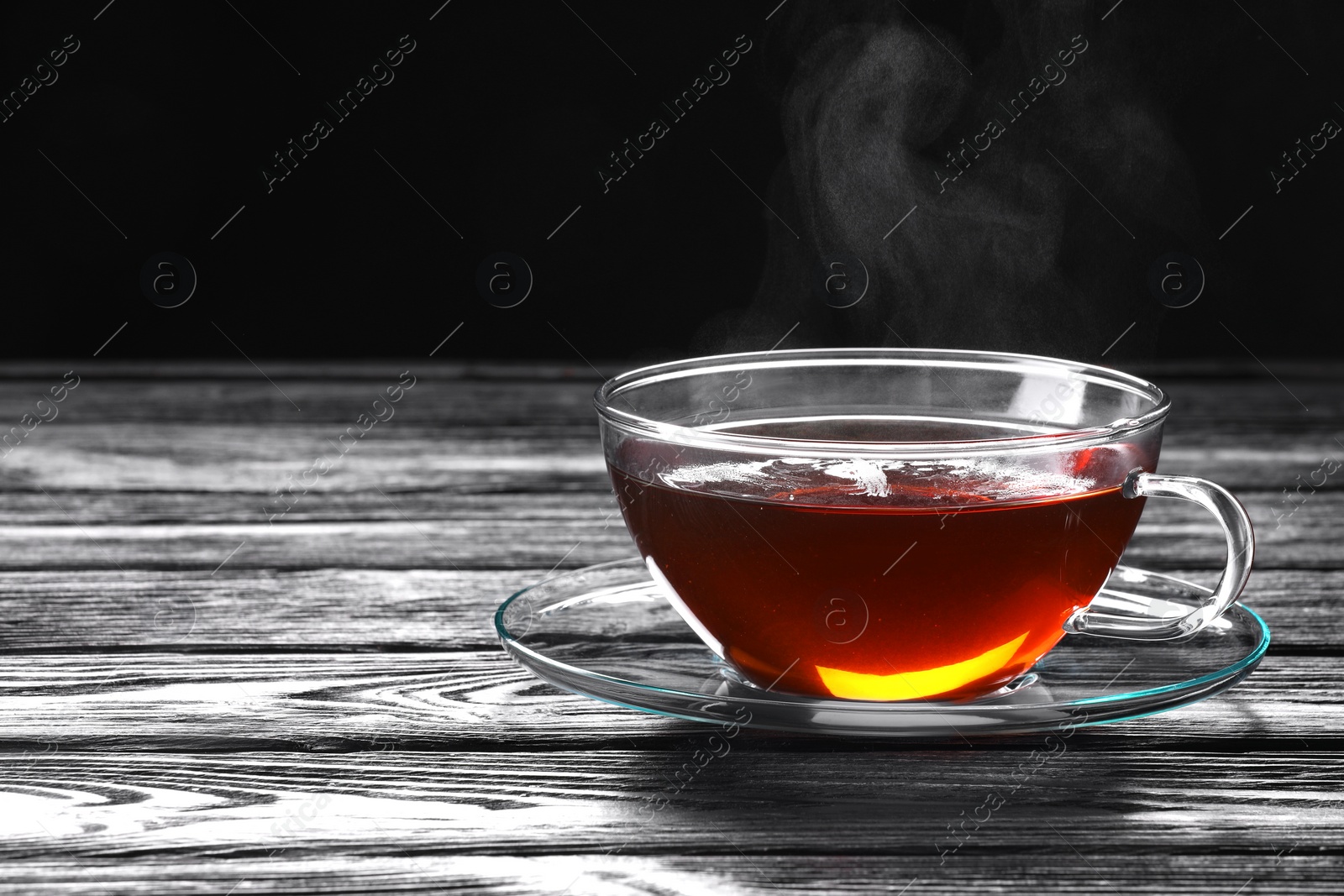 Photo of Glass cup of tea and saucer on wooden table against black background, space for text