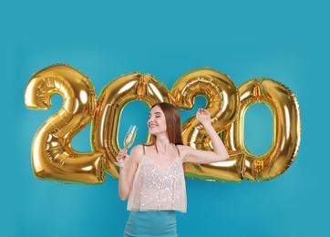 Happy young woman with glass of champagne near golden 2020 balloons on turquoise background. New Year celebration