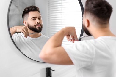 Photo of Handsome young man trimming beard with scissors near mirror in bathroom