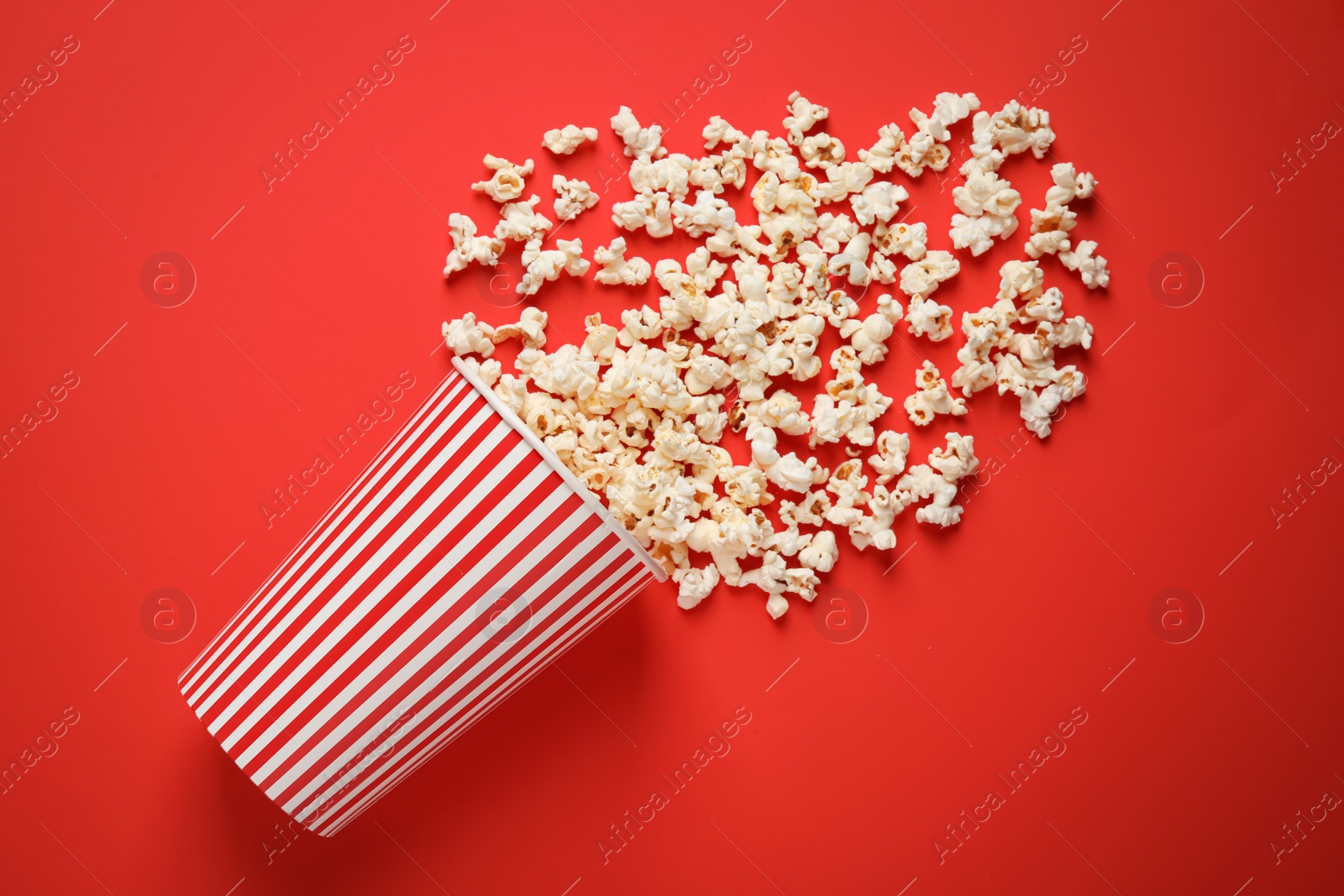 Photo of Overturned paper cup with delicious popcorn on red background, top view