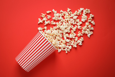 Photo of Overturned paper cup with delicious popcorn on red background, top view