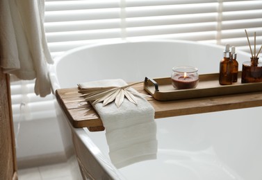 Photo of Wooden tray with cosmetic products, burning candle, reed air freshener and towel on bath tub in bathroom