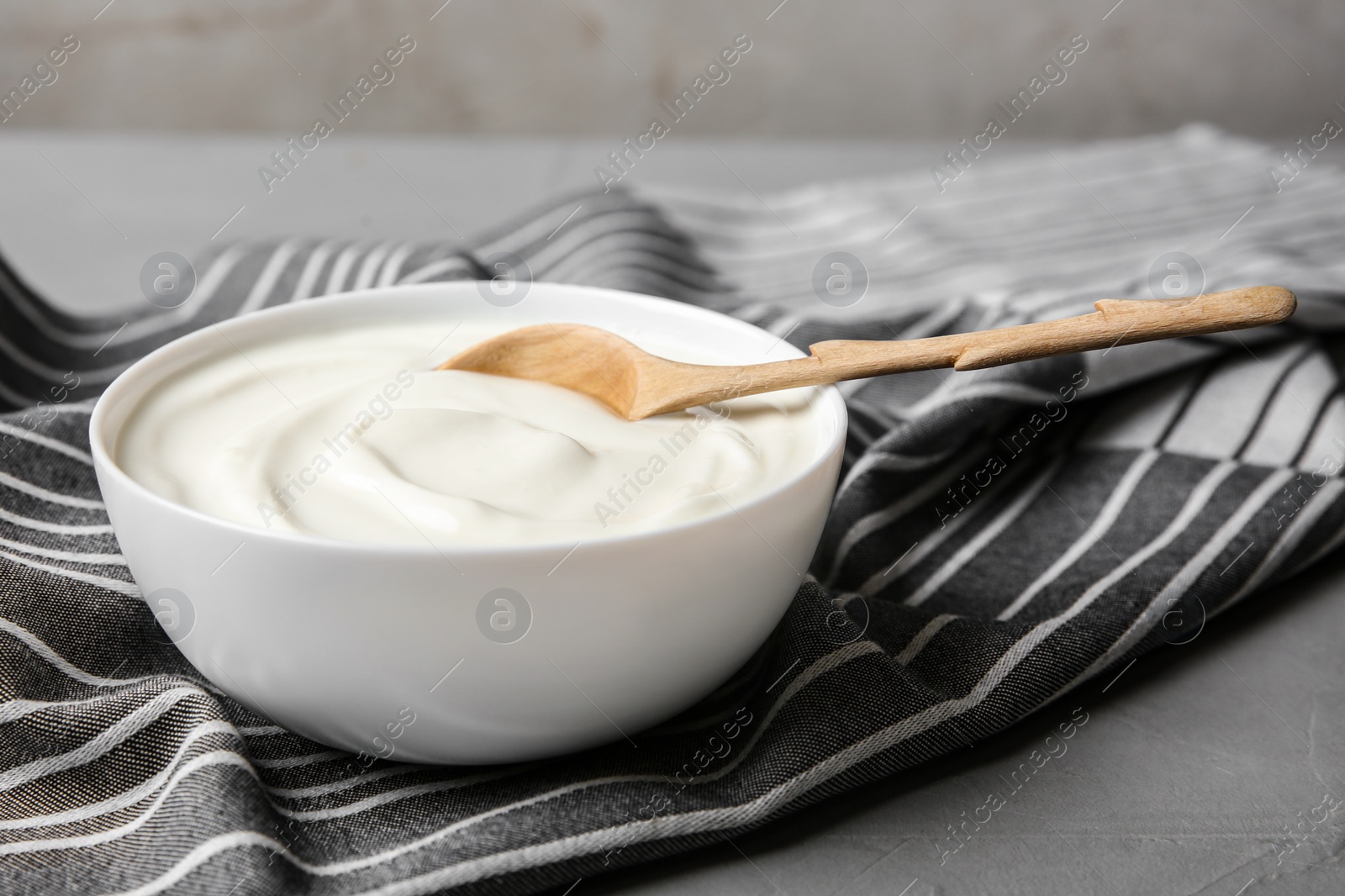 Photo of Bowl of sour cream with wooden spoon and napkin on grey marble table