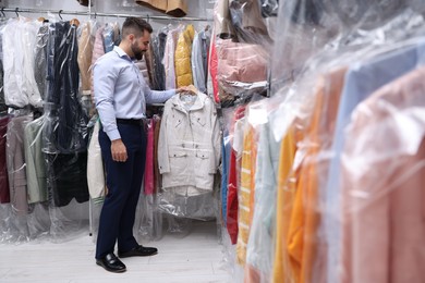 Photo of Dry-cleaning service. Happy worker holding hanger with jacket in plastic bag near other clothes indoors