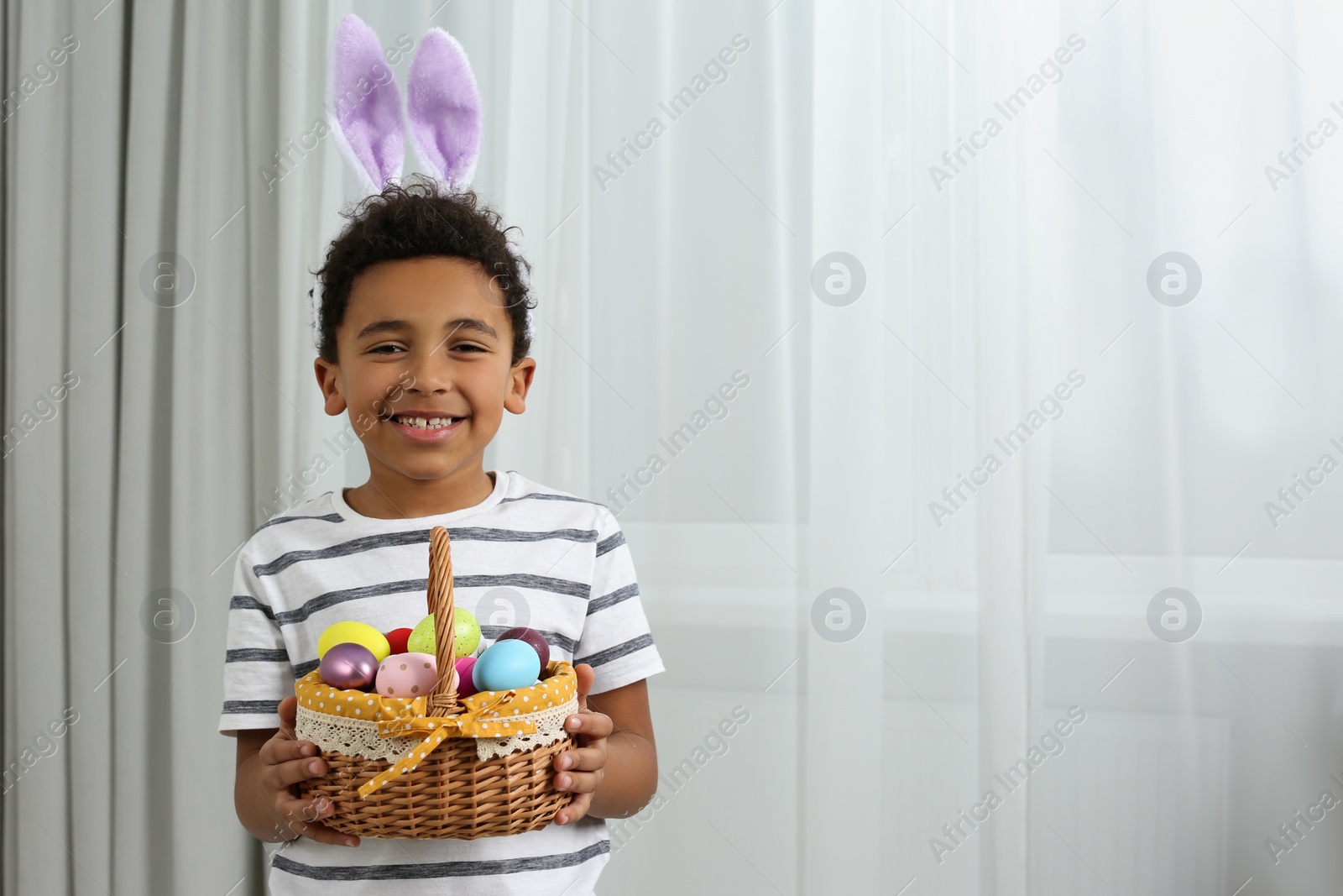 Photo of Cute African American boy in bunny ears headband holding wicker basket with Easter eggs indoors, space for text