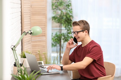 Handsome teenage boy talking on phone and using laptop at table in room