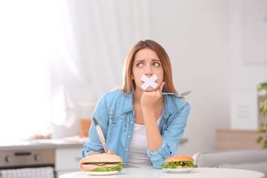 Photo of Sad young woman with taped mouth and burgers at table in kitchen. Healthy diet