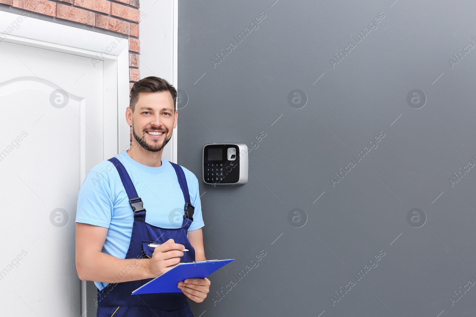 Photo of Male technician with clipboard near installed alarm system indoors