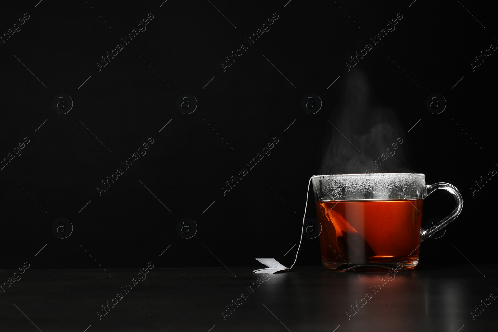 Photo of Glass cup of hot tea on table against black background, space for text