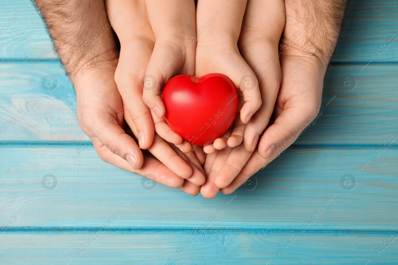 Photo of Parents and kid holding red heart in hands at light blue wooden table, top view