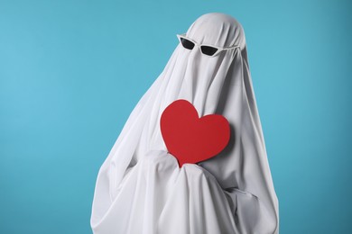 Cute ghost. Person covered with white sheet in sunglasses holding red heart on light blue background