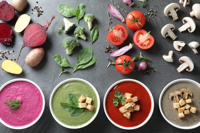 Photo of Different tasty cream soups in bowls and ingredients on dark grey table, flat lay