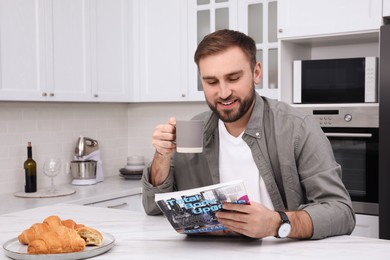 Photo of Handsome man reading magazine during breakfast at white marble table in kitchen