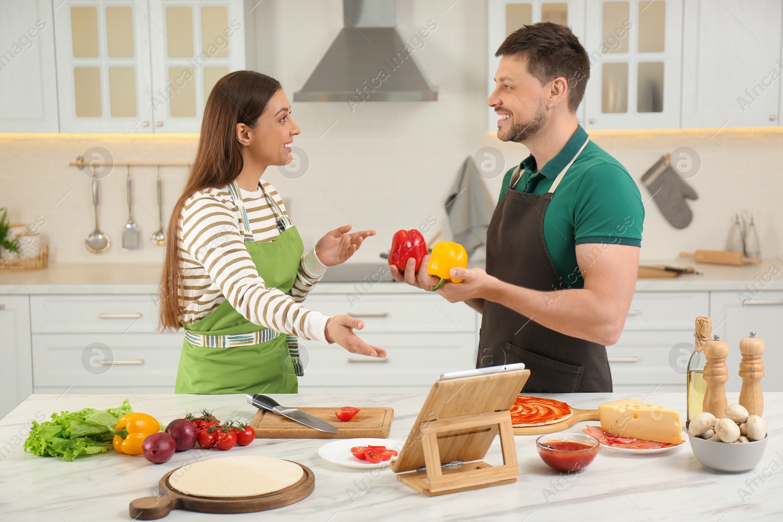 Photo of Happy couple making dinner together while watching online cooking course via tablet in kitchen