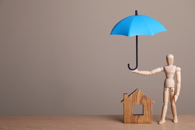 Mannequin holding small umbrella over house figure on wooden table. Space for text