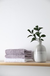 Photo of Stacked terry towels and green branches in vase on wooden shelf near white wall