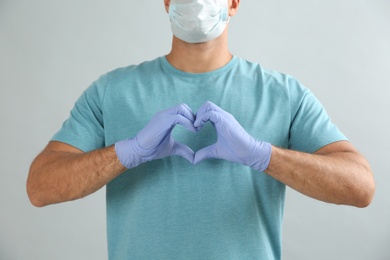 Man in protective face mask and medical gloves making heart with hands on grey background, closeup