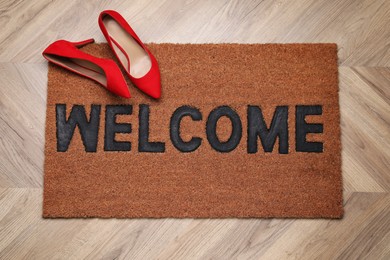 Stylish door mat with word Welcome and shoes on wooden floor, top view